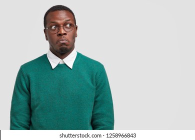 Puzzled dark skinned man looks thoughtfully and with bewilderment, focused aside, dressed in casual sweater, optical glasses, models over white studio wall with free space for your text or information