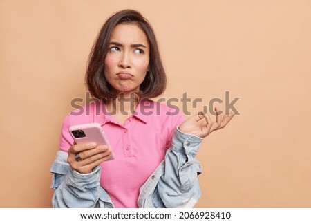Puzzled displeased beautiful Asian woman has sulking expression holds smartphone raises hand with bewilderment doesnt know how to download new application wears pink t shirt and denim jacket