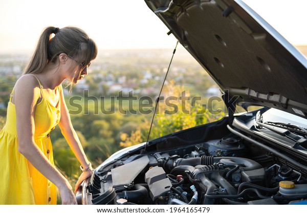 Puzzled female driver standing near her\
car with popped up hood looking at broken\
engine.