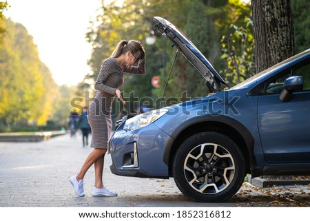Puzzled female driver standing on a city street near her car with popped up hood looking at broken engine.