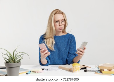 Puzzled Female Holds Two Modern Smart Phones, Downloads New Applications, Can`t Understand How To Use It, Has Puzzled And Tired Expression, Blows Cheeks In Bewilderment. People And Technology