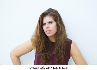 Puzzled Girl With Furrowed Brow Looking At Camera And Standing Against White Wall. Confused Young Woman Holding Hands On Hips. Misunderstanding Concept