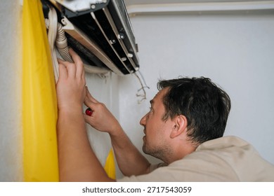 Puzzled HVAC professional disassembling wall-mounted air conditioning unit, using yellow drainage bag while carefully accessing electrical components and managing condensation water.