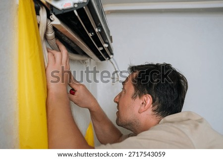 Puzzled HVAC professional disassembling wall-mounted air conditioning unit, using yellow drainage bag while carefully accessing electrical components and managing condensation water.