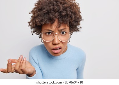 Puzzled indignant woman says so what raises hand and looks displeased at camera frowns face with dissatisfactionn wears big transparent glasses casual blue jumper isolated over white background