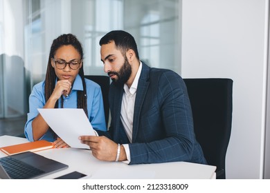 Puzzled male and female colleagues discussing information during cooperation briefing in office interior, multicultural partners in formal clothes analyzing paperwork reports and documents in company