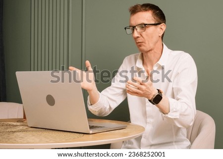 Puzzled mature human sitting at table and looking at laptop screen with misunderstood eyes. Dazed business man in glasses and white shirt working from home raised hands in shock.
