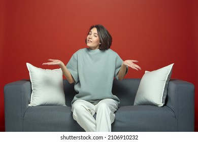 Puzzled Pretty Young Girl Dont Know Answer, Spread Hands, Shrug Shoulders, Sitting On Sofa On Red Studio Background