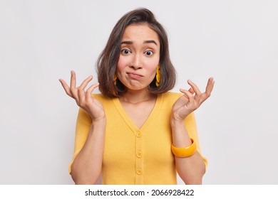 Puzzled questioned woman with dark hair clueless expression raises hands shrugs shoulders with bewilderment doesnt know how to act wears casual yellow jumper isolated over white studio background