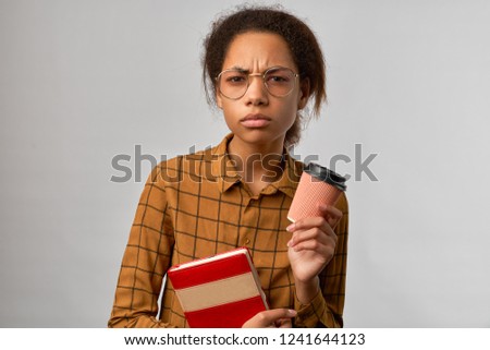 The puzzled student with a nervous indignant look on his face, holding a take-away coffee and a red textbook, isolated on a white background. A woman with dark skin looks intently into the camera. 