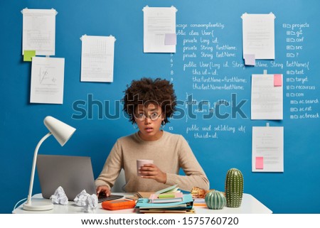 Puzzled surprised computer analyst has coffee break, completes report, reads presrelease published on web page, wears round spectacles, poses at desktop against blue wall with written information