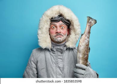 Puzzled surprised frozen man spends long hours outdoor during severe cold day dressed in grey winter jacket and gloves holds fish wears ski glasses isolated on blue background. Frosty weather
