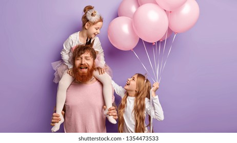 Puzzled Tired Busy Father Entertains Daughters, Feels Fatigue Of Playing Different Games On Party, Enjoy Birthday Celebration Stands Against Purple Background. Funny Sisters Look Happily At Each Other