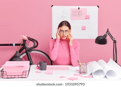 Puzzled tired female architect tries to complete working task develops planning project concentrated at papers poses at desktop. Displeased overworked woman graphic designer talks via mobile phone - Powered by Shutterstock