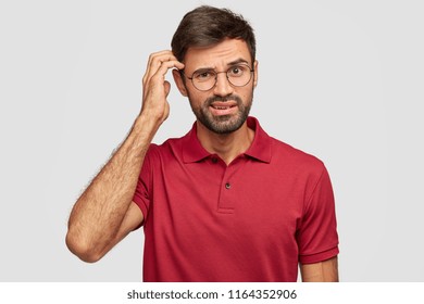 Puzzled unshaven guy scratches head, thinks deeply about something, has indignant look, dressed in casual clothes, stands against white background. Human facial expressions, emotions concept