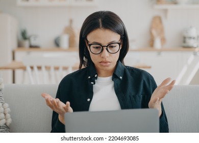 Puzzled Woman Working At Laptop Spreading Hands, Reading Email With Bad News At Home. Frustrated Female Freelancer In Glasses Looks At Screen, Shocked By Problems In Work, Error On Computer.