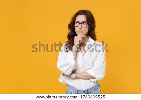 Puzzled young brunette business woman in white shirt glasses isolated on yellow background. Achievement career wealth business concept. Mock up copy space. Put hand prop up on chin, looking aside
