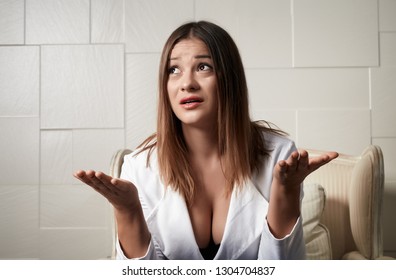 Puzzled young businesswoman with big breasts, seated on an armchair keeps hands raised in palms.