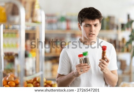 Puzzled young guy holding three spice bottles with different labels and lid, pondering options while shopping for piquant flavorful seasonings at grocery store