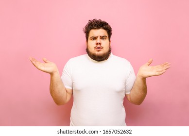 Puzzled young overweight man stands on a pink background and spreads his arms out to the sides. Confused fat guy does not know, surprise looking at the camera, isolated. Copy space