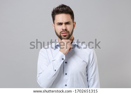 Puzzled young unshaven business man in light shirt posing isolated on grey wall background studio portrait. Achievement career wealth business concept. Mock up copy space. Put hand prop up on chin
