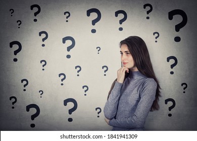 Puzzled Young Woman Has Multiple Questions, Keeps Hand Under Chin, Thoughtful Gesture, Looking Ahead Focused. Confused Student Girl And Different Interrogation Marks. Unsure Teen Thinking For Answer. 