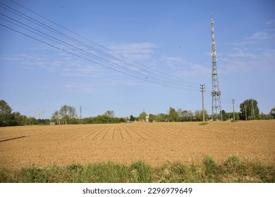 Pylons  and overhead cables in a ploughed field with a radio mast in the distance on a sunny day in the italian countryside