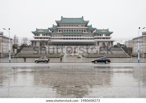 Pyongyang, North Korea, April 2019 - Cars\
drive past the Grand People\'s Study House and Kim Il Sung Square on\
a rainy day in\
Pyongyang.