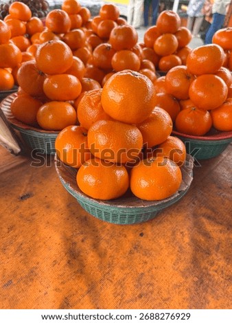 A pyramid stack of fresh, ripe Mandarin Oranges (Tangerines) displayed in a green basket at a bustling local outdoor market. A perfect, bright image for Vitamin C, healthy eating, and citrus fruit.