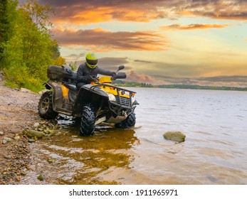 Quad bike on the river bank. The ATV driver drove into water. Quad bike on the background sunset. A man travels on a quad bike. A yellow ATV stands in lake. Concept - outdoor activity.