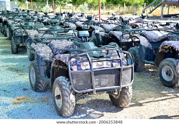 Quad bikes lined up in several\
rows on the station for rent. Cars are in the shade of an\
awning.
