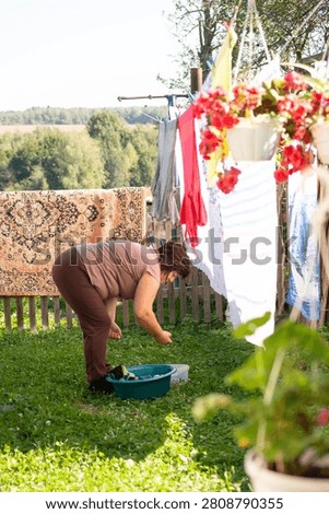 In a quaint backyard, an individual hand-washes laundry next to a clothesline. The setting has a carpet hanging, flower pots, and a view of a rural landscape, creating a domestic scene.