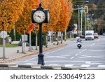 A quaint street at Harrison Lake, BC, featuring a vintage clock and vibrant autumn leaves. A motorcycle rider adds dynamic movement to the scene under a clear sky.