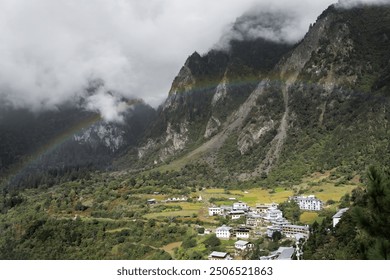 A quaint village nestled amidst lush, misty mountains, with a vibrant rainbow arching gracefully across the cloudy sky. - Powered by Shutterstock