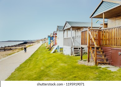 Quaint Wooden Beach Huts In A Row Along A Grass Verge In Front Of The Promenade That A Man Is Cycling Along, By The Beach And Sea In Tankerton, Whitstable, Kent, UK