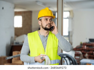 Qualified builder working on a construction site indoors is talking on a mobile phone, discussing important work issues