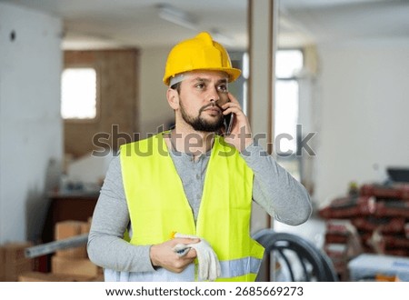 Qualified builder working on a construction site indoors is talking on a mobile phone, discussing important work issues