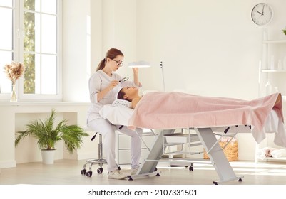 Qualified cosmetologist looking at patient's face under loupe and lamp magnifier. Licensed dermatologist or beautician examining lady's facial skin before doing professional cosmetic treatment