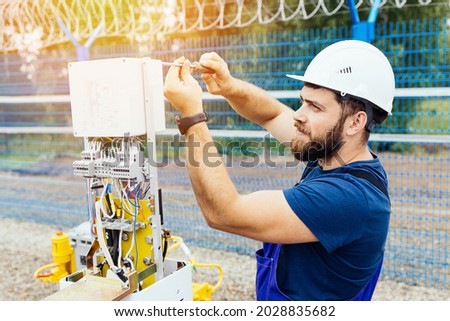 A qualified electrical engineer sets up a control and measuring device at an industrial facility on a summer day. Specialist power engineer in helmet and uniform at work. An authentic portrait.