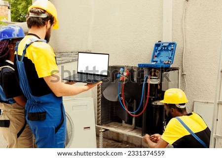 Qualified electrician cleaning and lubricating outside air conditioner internal compressor, checking freon level while african american colleague checks maintenance schedule on laptop