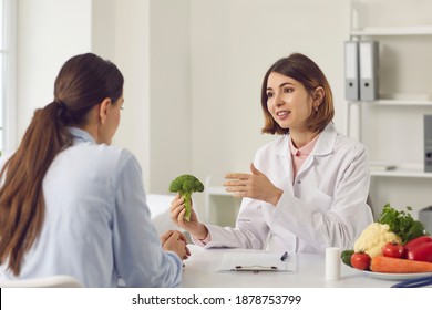 Qualified Female Dietitian, Nutritionist, Healthy Food Expert, Doctor Of Alternative Medicine, Consultant In Health Center Holding Broccoli And Telling Young Woman About Benefits Of Eating Vegetables