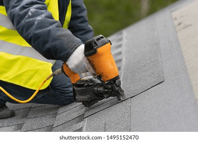 Qualified Roofer Worker In Protective Uniform Using Air Or Pneumatic Nail Gun And Installing Asphalt Or Bitumen Shingle On Top Of The New Roof Under Construction Residential Building