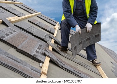 Qualified roofer worker in uniform work wear holding in hands and installing asphalt or bitumen tile on top of the roof under construction house against beautiful blue sky on background