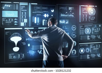 Qualified specialist. Calm clever experienced programmer standing with his hand resting on his hip and thoughtfully looking at the information on a transparent screen