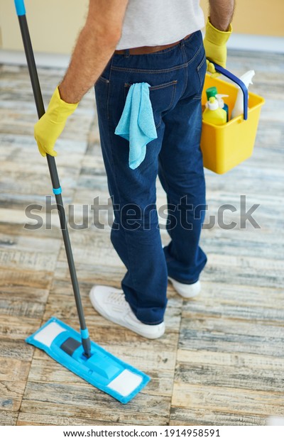 Quality cleaning. Professional male cleaner\
holding mop and plastic bucket with detergents, ready for cleaning\
floor in the kitchen
