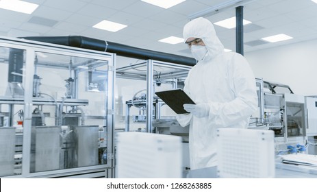 Quality Control Check: Scientist Using Digital Tablet Computer And Wearing Protective Suit Walks Through Manufacturing Laboratory. Product Manufacturing: Pharmaceutics, Semiconductors, Biotechnology.