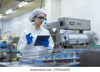 Quality control technician using tablet in canned fish factory, inspecting products on an automated production line with focus on food safety, hygiene standards, packaging, manufacturing.