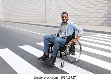 Quality Of Life And Impairment Concept. Positive Disabled Black Guy In Wheelchair Crossing City Street. Handicapped Young Man Going For Walk Downtown Area, Using Crosswalk Alone