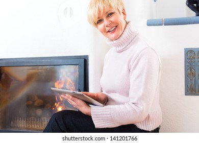 Quality Of Life - Older Woman Or Female Pensioner Sitting At Home In Front Of The Furnace, Writing Emails On The Tablet Computer Or Reading A E-book