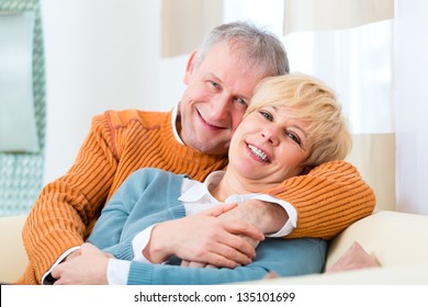 Quality Of Life - Two Elderly People Sitting At Home On The Couch, He Embraces His Wife